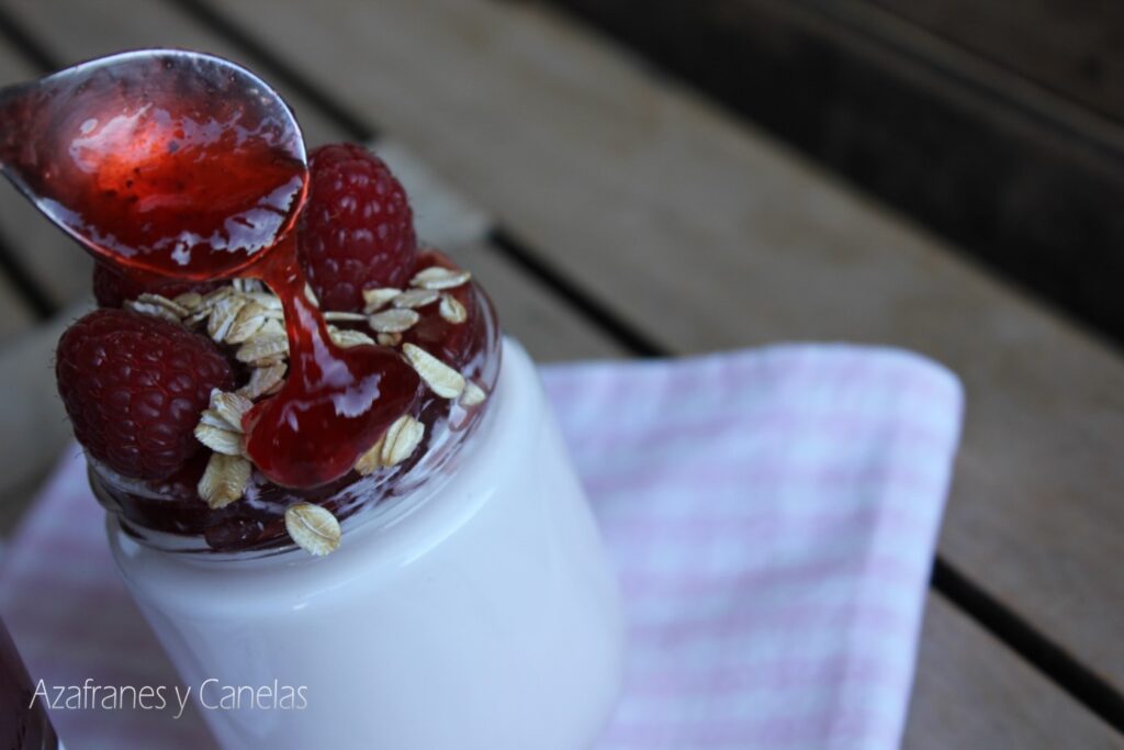 Como hacer yogurt casero. Vaso de yogurt blanco con frambuesas y copos de avena. Una cucharada de mermelada chorrea sobre la fruta.