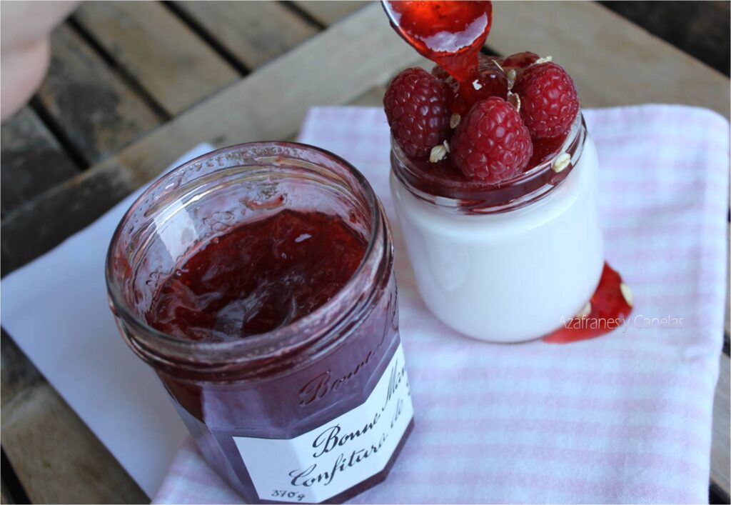 Como hacer yogurt casero. Vaso de yogurt blanco con frambuesas y copos de avena. Una cucharada de mermelada chorrea sobre la fruta. Se ve el bote de mermelada rojo intenso.